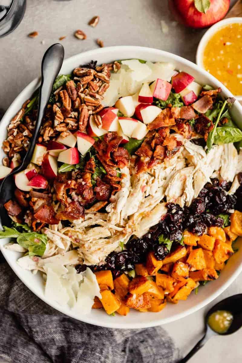 large white bowl filled with dark lettuce greens topped with pecans, apples, Manchego cheese, bacon, chicken, cranberries and squash