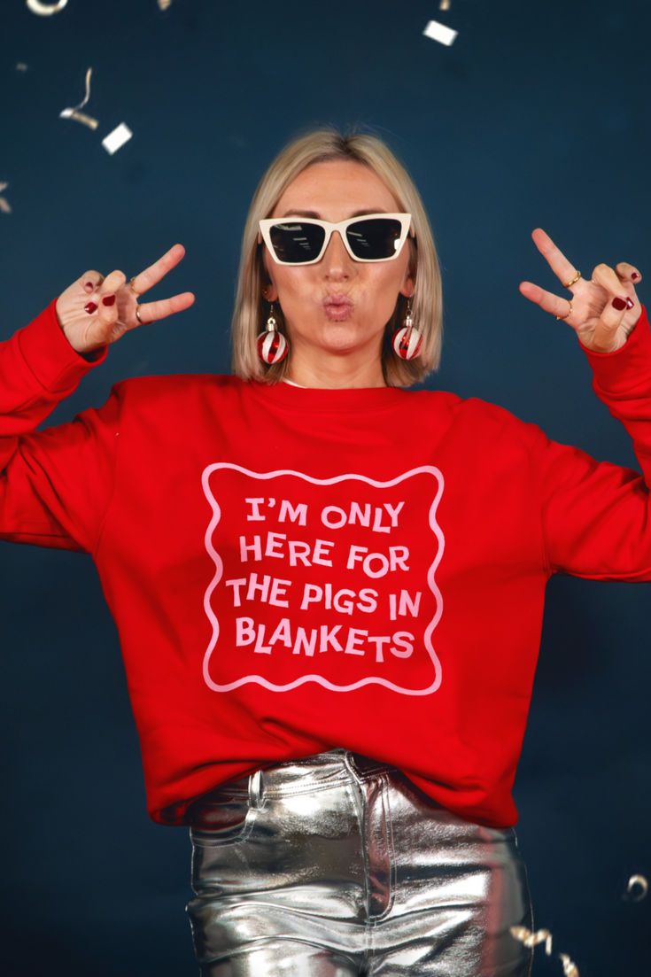 This contains: woman stood in front of navy wall wearing a bright red and pink sweatshirt with the slogan "I'm only here for the pigs in blankets"