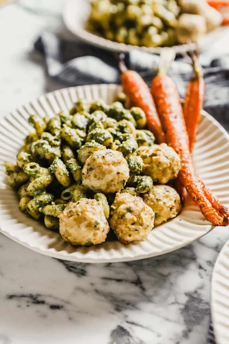 Photograph of chicken meatballs in a white bowl with pesto pasta and roasted carrots
