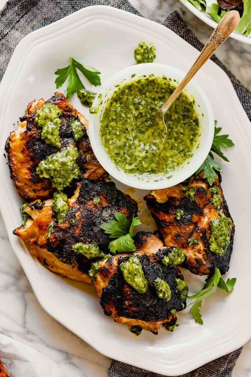 Grilled chicken thighs on a white plate drizzled with chimichurri sauce with a bowl of green sauce off to the side