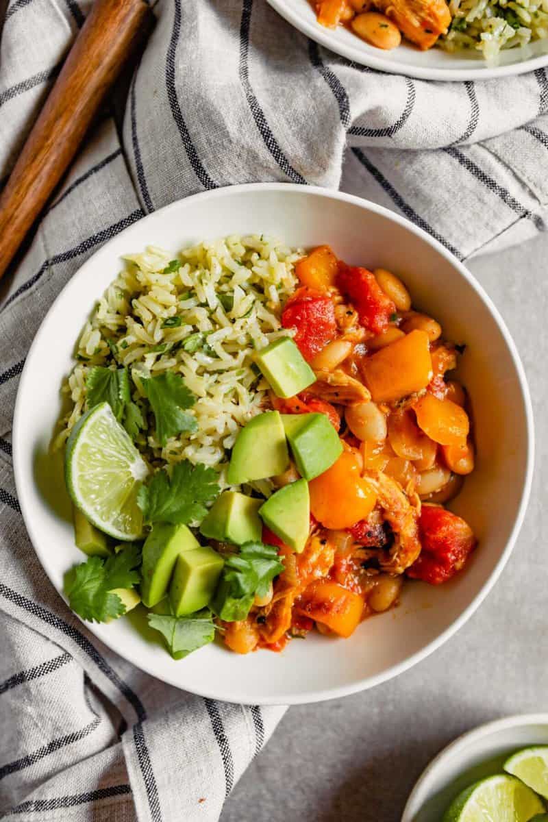 Overhead image of a while bowl filled with brown rice, a saucy veggie mixture, diced avocado and lime wedges.