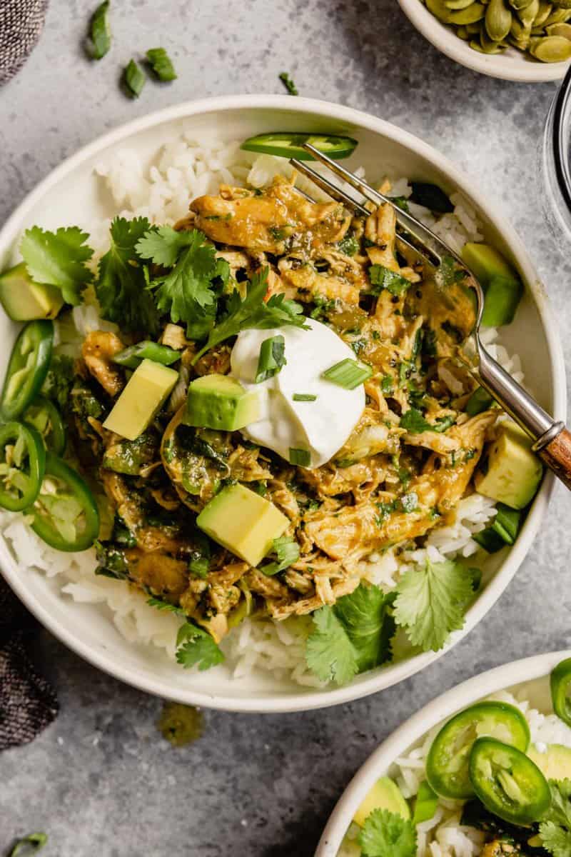 shredded salsa verde chicken in top of white rice in a white bowl garnished with sour cream, avocado, scallions, and cilantro